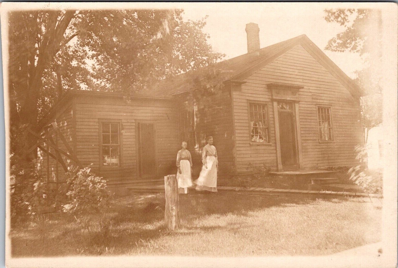 RPPC Two women wearing aprons in front of wood house tree stump